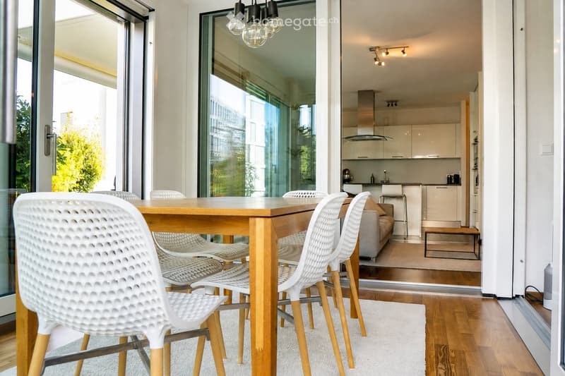 Bright, modern dining area with a wooden dining table and white chairs, leading to a sleek kitchen with white cabinets and stainless steel appliances.