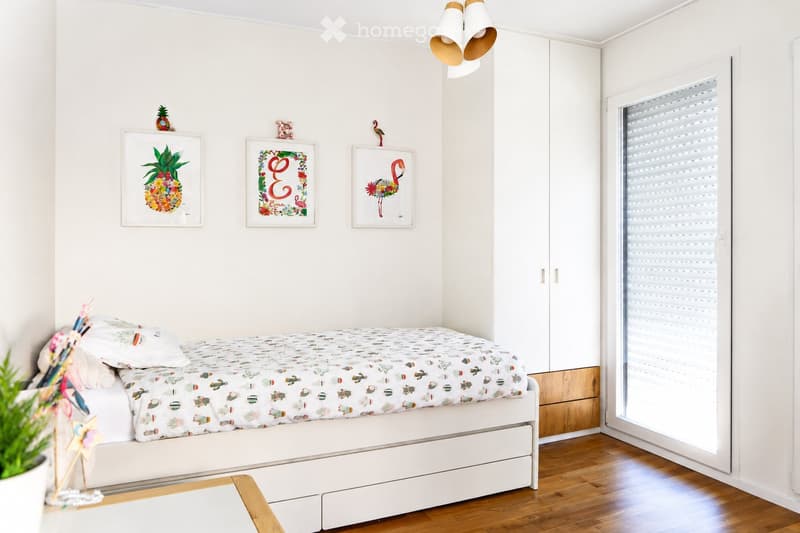A modern, minimalist bedroom with a white bed, colorful bedding, a built-in wardrobe, and decorative wall art.