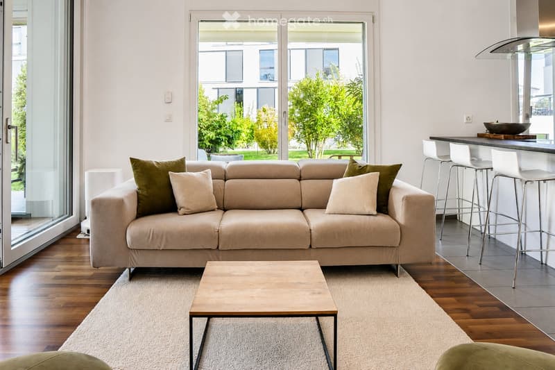 Modern living room with a beige sofa, wooden coffee table, white bar counter with stools, and large windows overlooking a green garden.