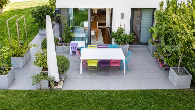 A modern outdoor patio with a white table, colorful chairs, and potted plants, connected to a house with large glass doors.