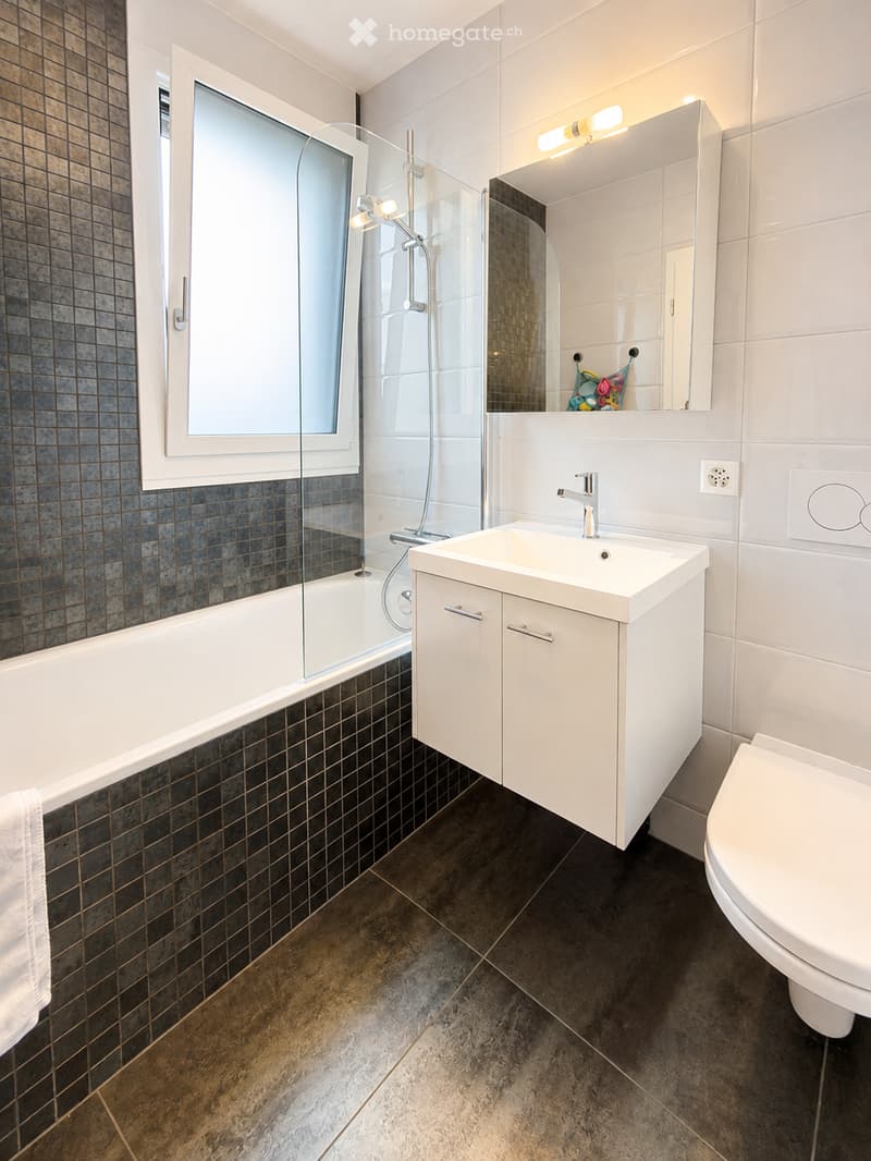 Modern bathroom with a bathtub, sink, toilet, and a large mirror above the sink. The walls are tiled in dark and light shades, and the floor is made of dark stone tiles.