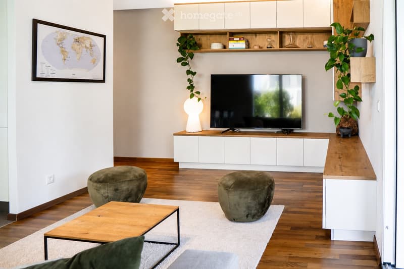 Modern living room with wooden flooring, white walls, and a neutral color palette. Features a television on a white cabinet, two green ottomans, a wooden coffee table, a floor lamp, and built-in shelving with decorative items and plants.