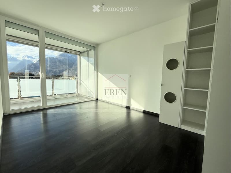 Empty room with dark wooden flooring, white walls, large glass windows and sliding doors leading to a balcony with a scenic mountain view.