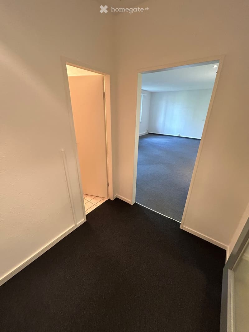 Empty hallway with beige walls, black carpet, and two open doors leading to other rooms.