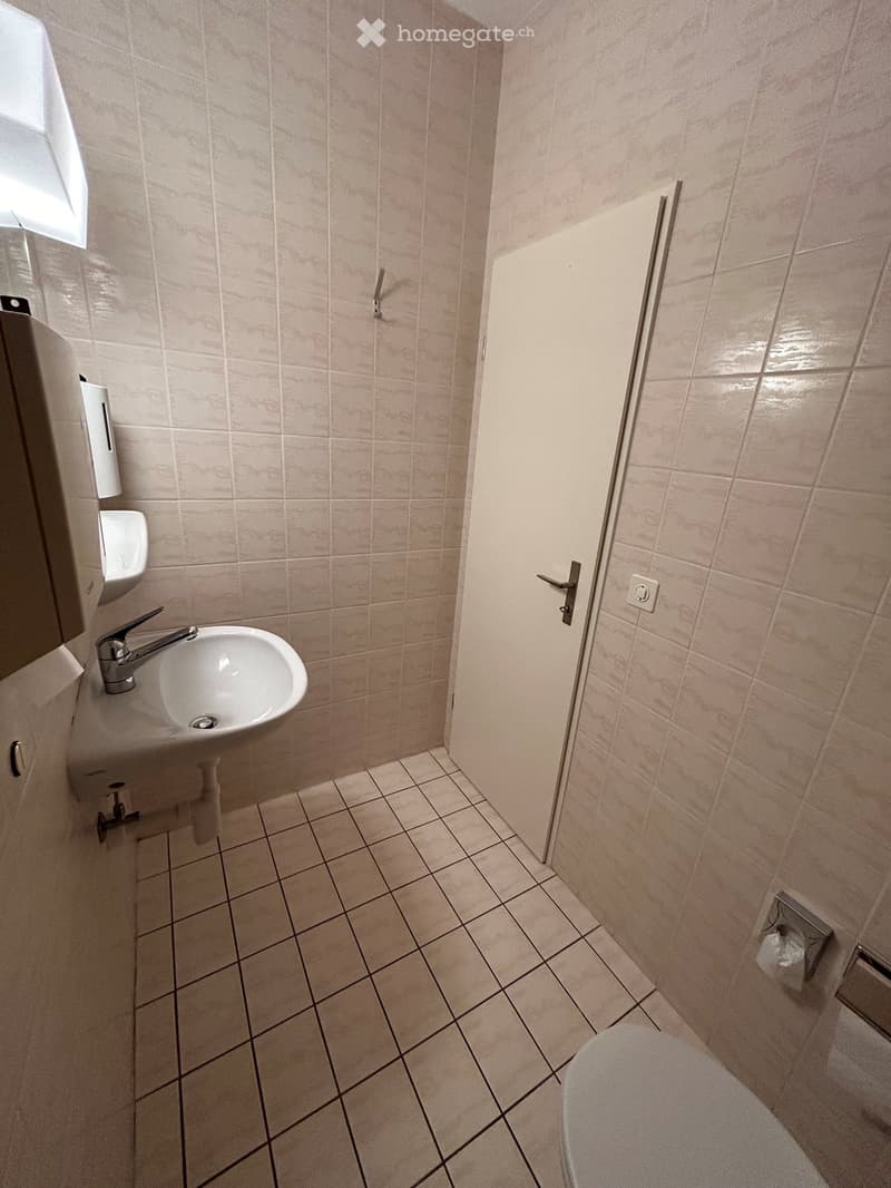 A small bathroom with tiled walls and floor, featuring a sink with a faucet, a toilet, and a door.