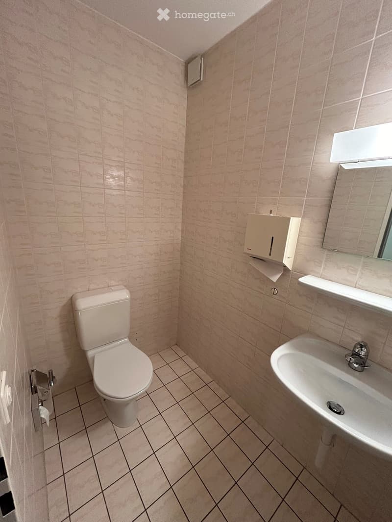 Small bathroom with beige tiled walls and floor, featuring a toilet, sink with faucet, mirror, and paper towel dispenser.