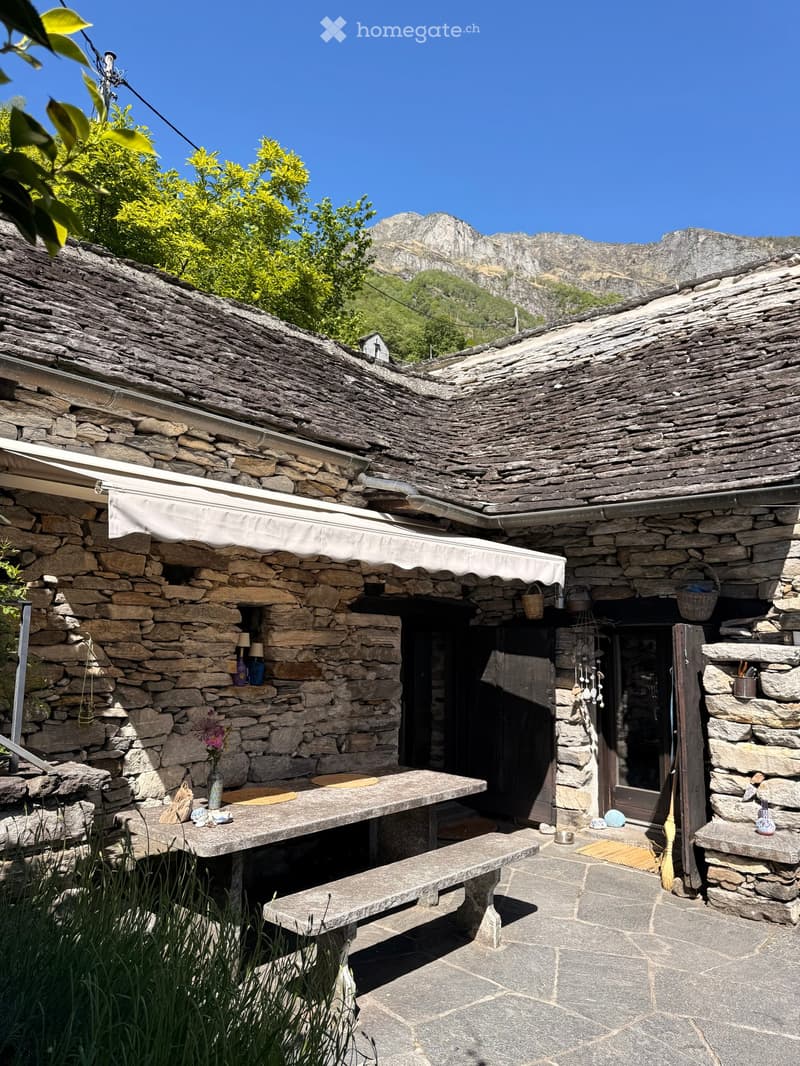 Stone outdoor seating area with a table, bench, vase with flowers, and baskets, set against a stone building with a view of mountains.