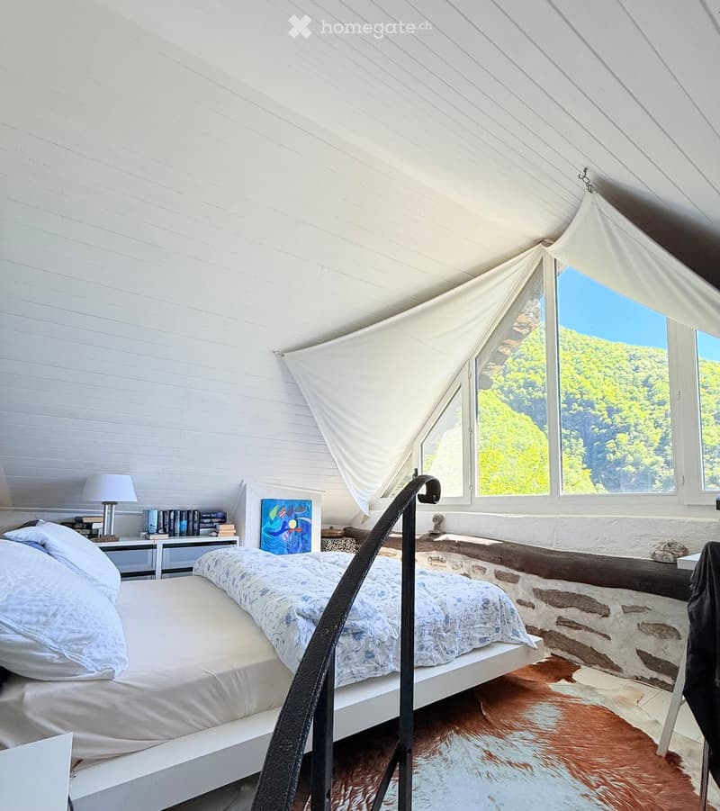 Bright bedroom with a large bed, nightstand, lamp, books, and a painting, featuring large windows with a scenic view.