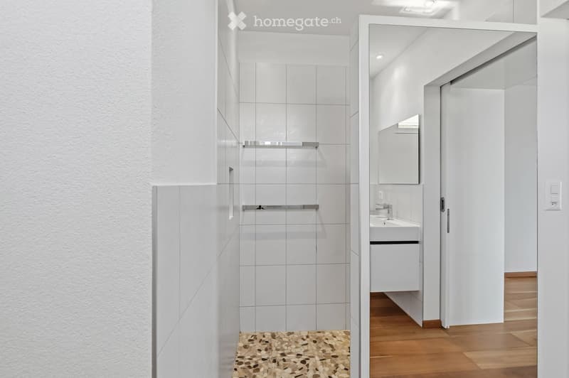 Modern bathroom with white tiles, a glass-enclosed shower, a sink with a mirror, and a wooden floor.