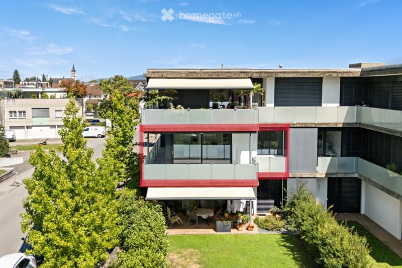 Modern multi-story building with red and white facade, large windows, balconies, and surrounding greenery.