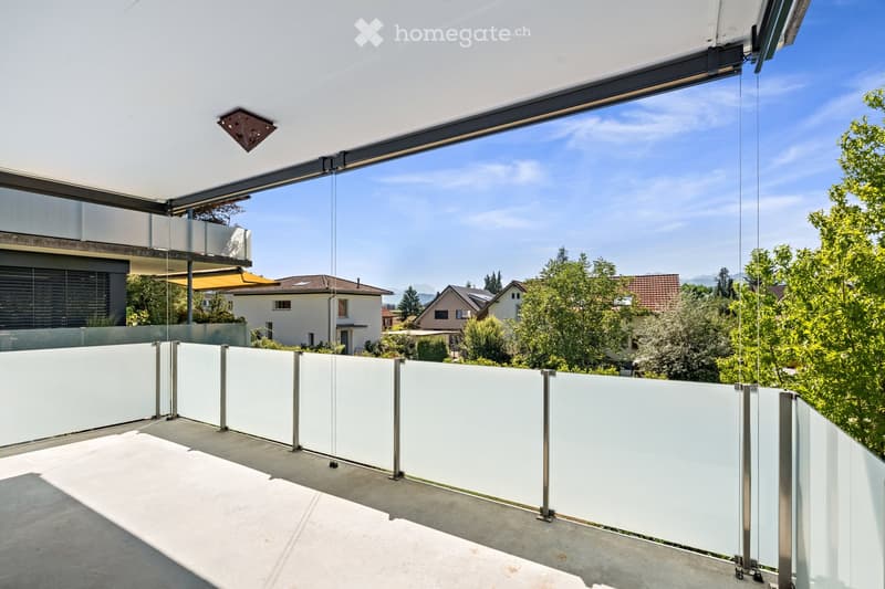 Modern balcony with glass railings, concrete floor, and a retractable roof, overlooking a residential neighborhood with trees and houses.
