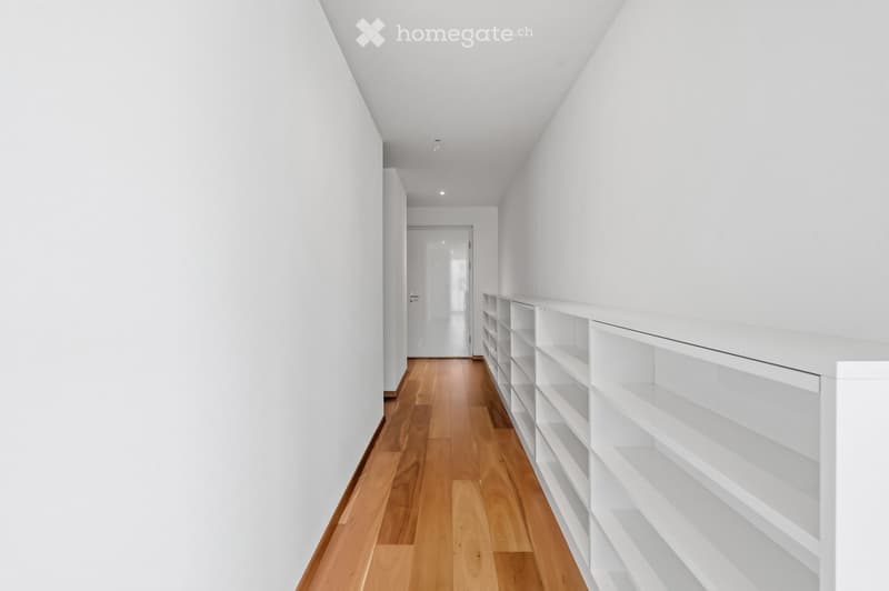 A clean, modern hallway with white walls, wooden flooring, and built-in white shelving units on one side.