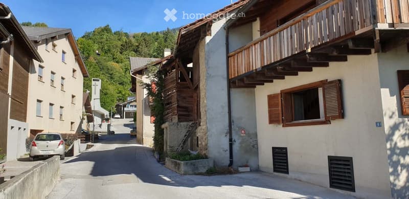 A row of traditional wooden and stone houses in a narrow village street, surrounded by greenery and mountains.