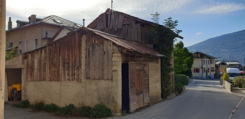 A rustic wooden building with a stone base and a corrugated metal roof, situated on a narrow street with other residential structures and a mountainous backdrop.