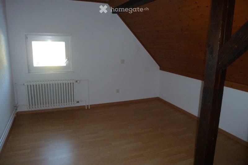Empty attic room with wooden floor, white walls, and a single window letting in natural light. A radiator is mounted on the wall below the window.