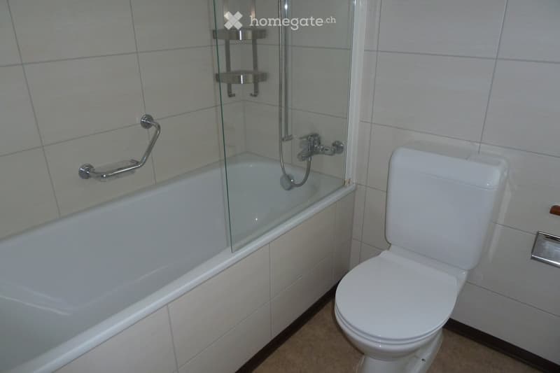 Modern bathroom with a white bathtub, glass shower enclosure, and a toilet. Grab bars are installed for safety, and the walls are tiled in light gray.