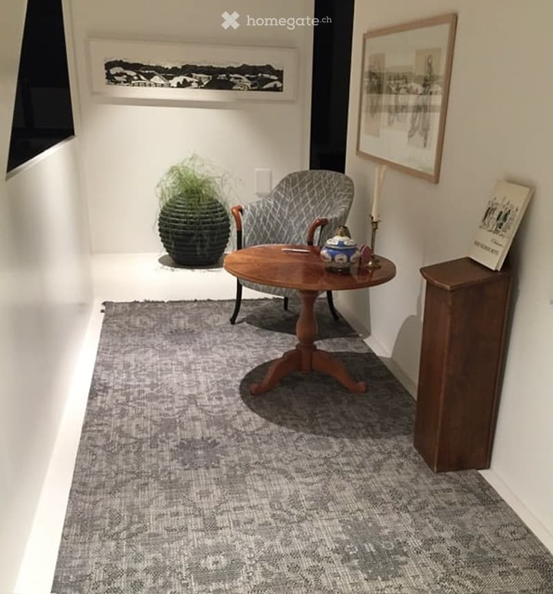 A small, well-lit room with a grey armchair, a wooden coffee table, a grey rug, a decorative plant, and a wooden cabinet with a book on top.