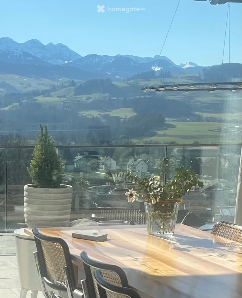 Balcony with a wooden dining table, chairs, a flower vase, and a potted plant, offering a scenic mountain view.