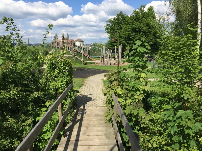 A wooden pathway lined with wooden railings leads through a lush, green garden area with dense foliage on both sides. In the background, a playground with climbing structures and a slide is visible.