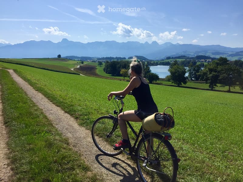 A woman rides a bicycle on a dirt path through a lush green field with a scenic mountain backdrop and a lake in the distance.