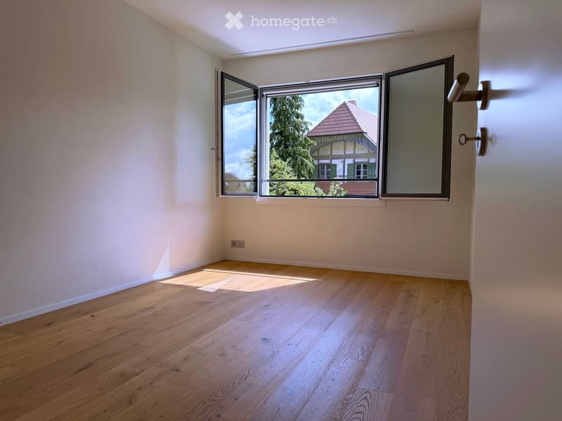 Empty room with light wooden flooring and large windows offering a view of a house and trees outside.