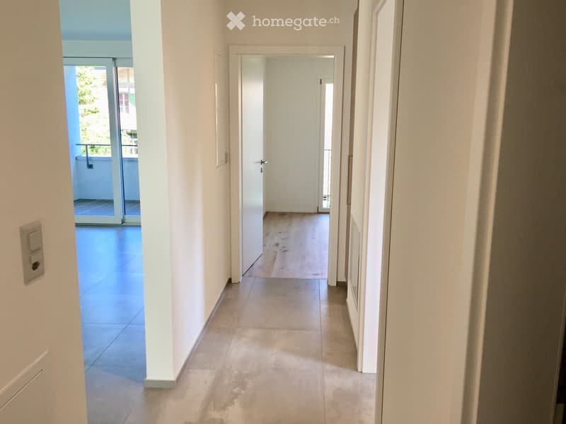 Empty hallway with white walls, tiled floor, and a door leading to a balcony area with wooden flooring.