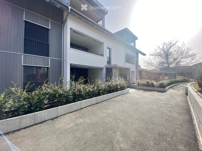 Modern two-story building with white and grey exterior, large windows, balcony, and small garden area.