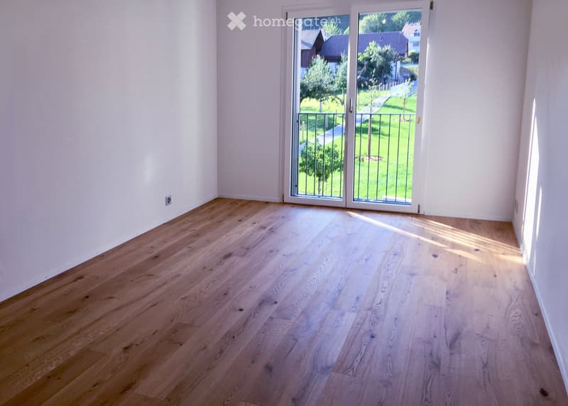 Empty room with light wooden flooring and white walls, featuring large glass doors leading to a green outdoor area.