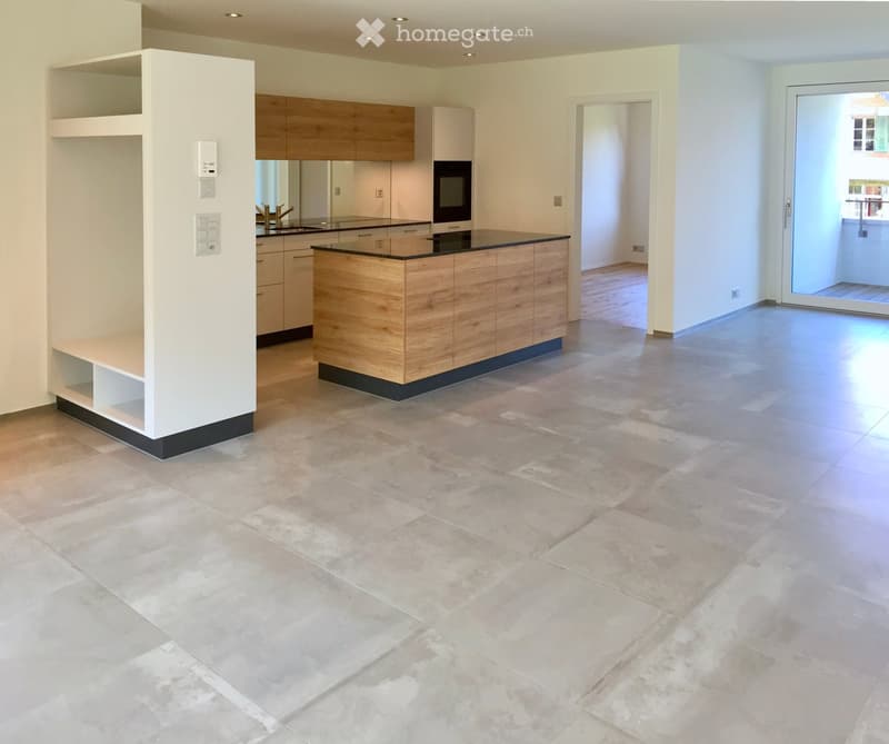 Modern kitchen with light wooden cabinets, a central island, and a large open space. The floor is tiled in a light beige color, and there is a sliding glass door leading to another room.
