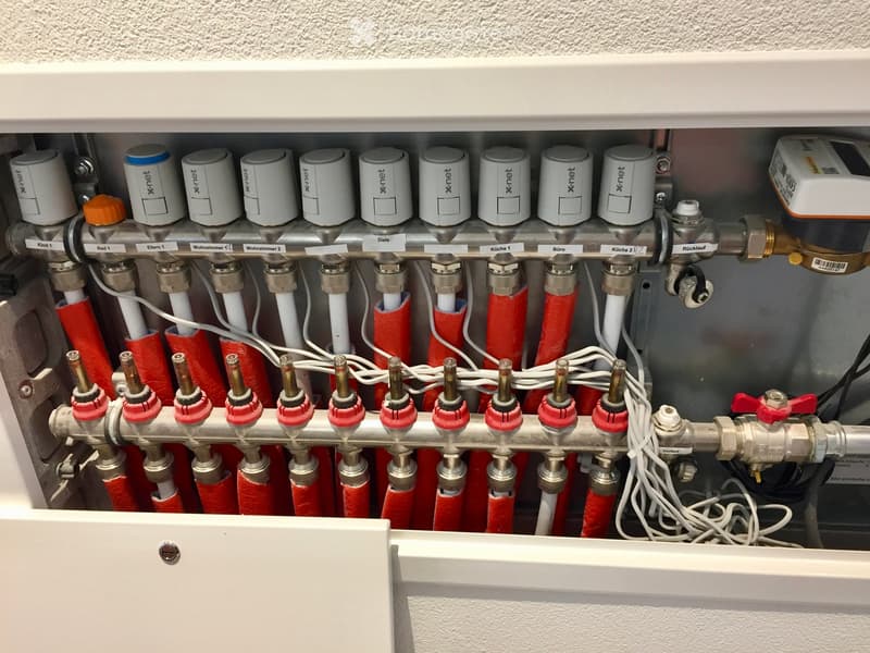 A close-up view of a heating system control panel with multiple valves, pipes, and thermostats labeled for different rooms.