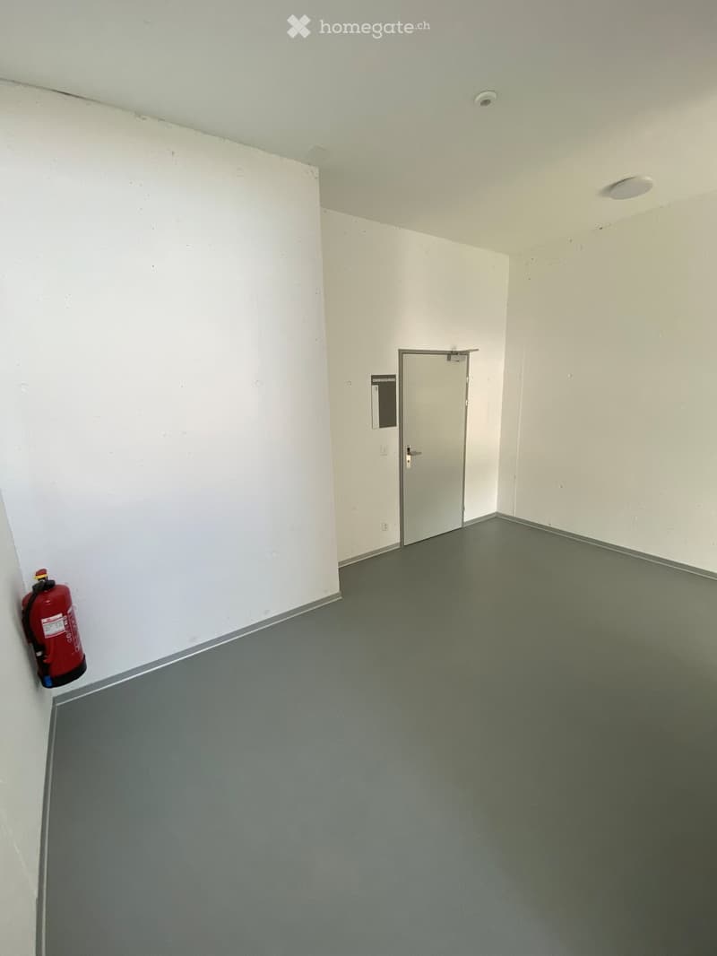Empty room with white walls, grey floor, and a single door leading to another space. A red fire extinguisher is mounted on the wall.