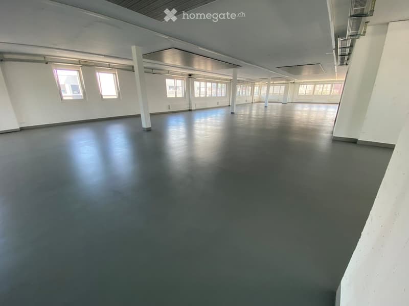 Empty room with white walls, large windows, and a dark grey floor. No furniture or equipment is present.