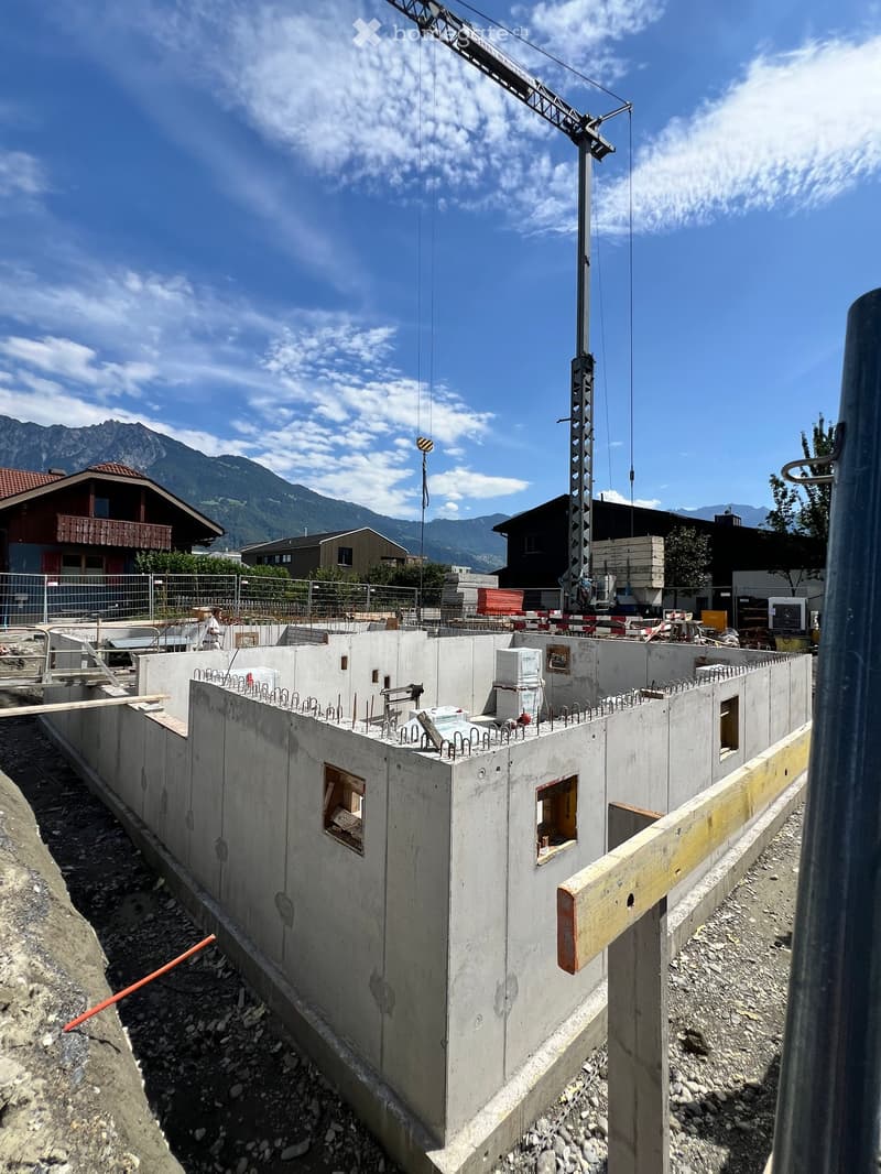 A partially constructed building with concrete walls, exposed rebar, and a crane in the background, set against a mountainous backdrop.