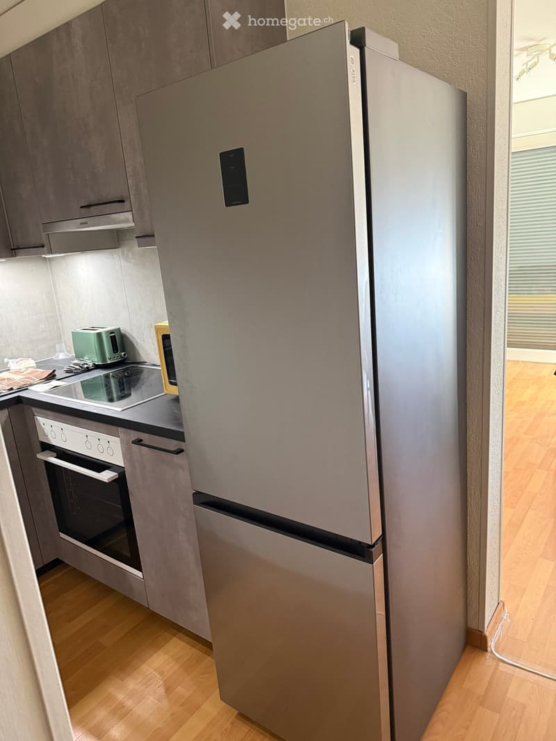 Modern kitchen with stainless steel appliances, including a refrigerator, oven, and toaster, set against grey cabinets and a wooden floor.