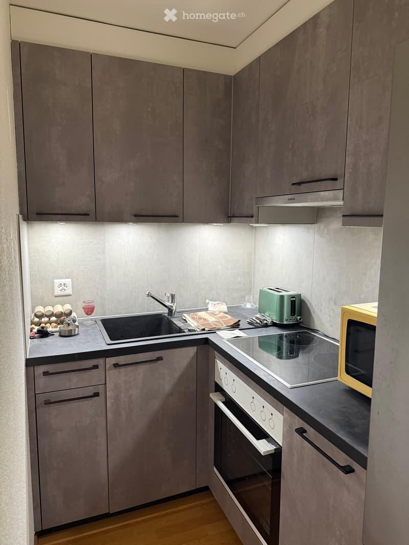 Modern kitchen with grey cabinets, stainless steel sink, built-in oven, microwave, toaster, wine rack, and wine glass on the counter.