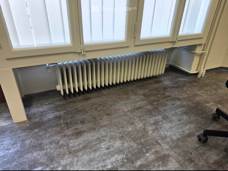 Empty room with a radiator under a window and a worn, stained concrete floor. No furniture or equipment is visible.
