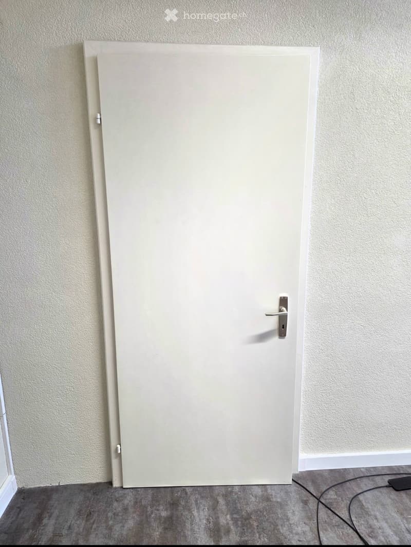 Empty room with a closed white door, light-colored walls, and a grey concrete floor. No furniture or equipment is visible.