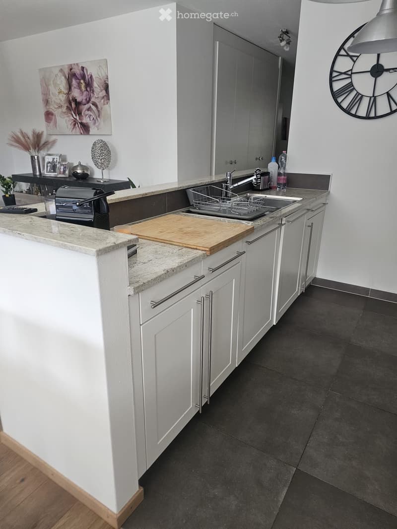 Modern kitchen with white cabinets, granite countertops, and stainless steel sink. Features a wooden cutting board, clock on wall, and decorative items on a side table.