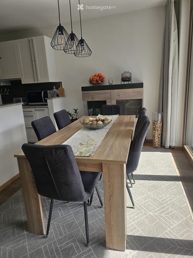 Modern dining area with a wooden dining table, six dark grey chairs, a decorative bowl with decorative items, a fireplace, and pendant lights. The area is connected to a kitchen with white cabinets and a microwave.