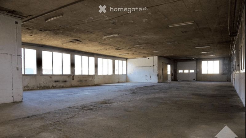 Empty industrial-style room with concrete floors and walls, large windows, and exposed ceiling beams. No furniture or equipment present.