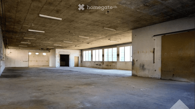 Empty industrial-style room with concrete floors and walls, large windows, and exposed ceiling with fluorescent lighting.