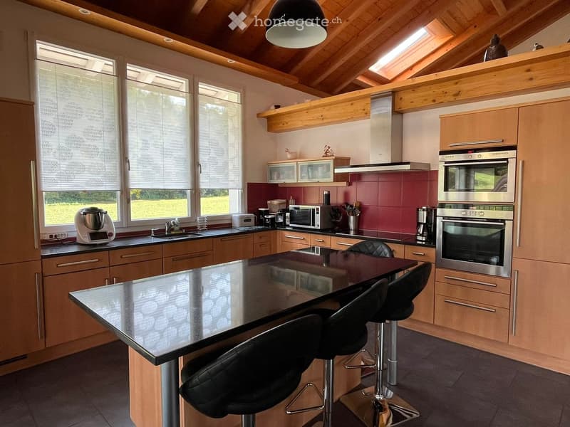 Modern kitchen with wooden cabinets, stainless steel appliances, a central island with bar stools, and large windows with blinds.