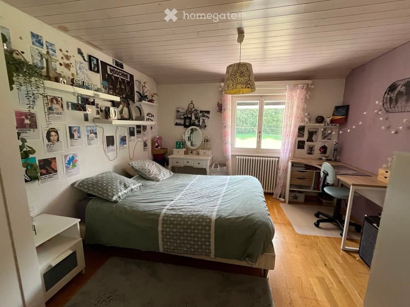 Cozy bedroom with a double bed, desk, chair, mirror, shelves, and a hanging lamp. The room features wooden flooring, a window with curtains, and various decorations including posters and a plant.