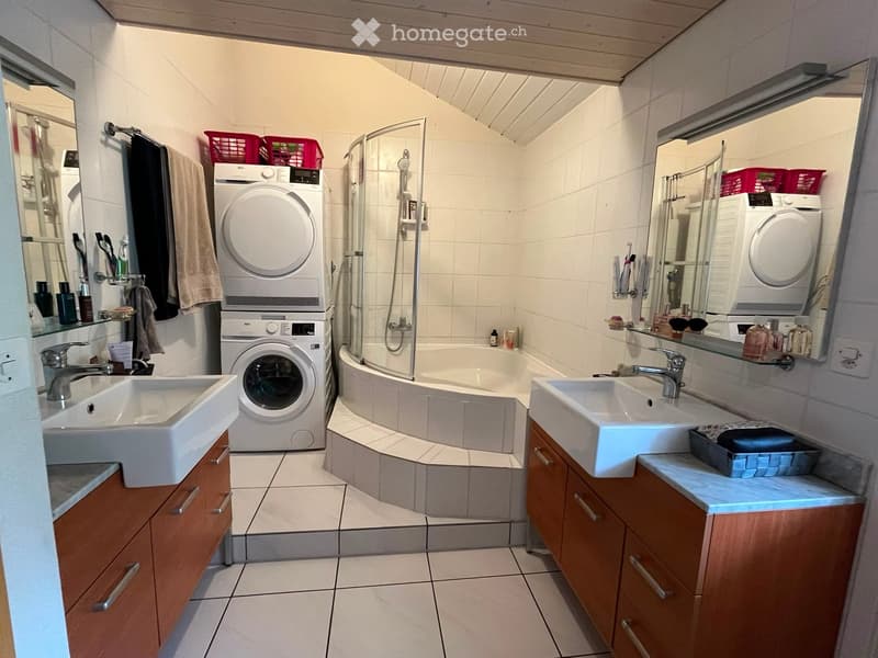Modern bathroom with two sinks, a bathtub with a glass shower enclosure, a washing machine, and wooden cabinets.