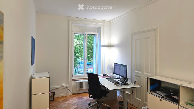 A modern, well-lit office space with a large window, a desk with a computer setup, a chair, a fan, and a cabinet with a printer.