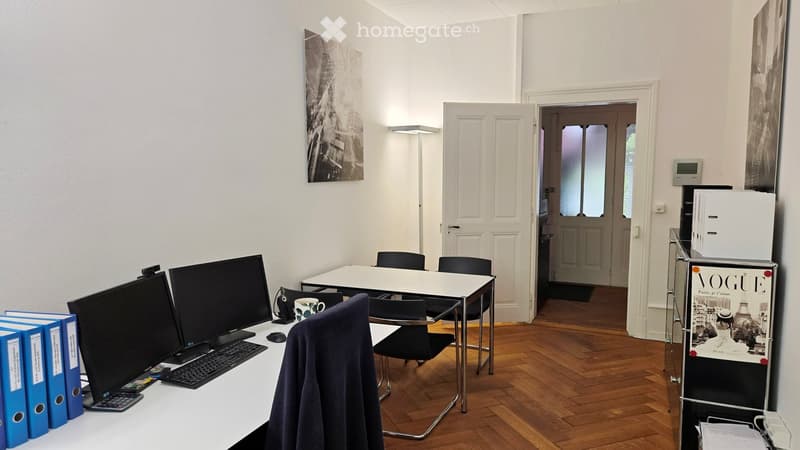 Office space with wooden flooring, white walls, a table with chairs, two computer screens, keyboard, mouse, a cup, and a poster on the wall.