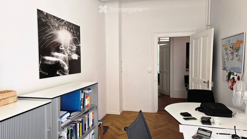 A modern office space with a wooden floor, white walls, and large windows. The room features a desk with a chair, a bookshelf filled with books, a fan, and a table with office supplies including a calculator, a cup, and a backpack.