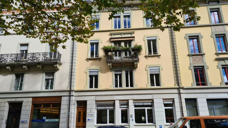 Multi-story residential building with light yellow facade, multiple windows, balconies with railings, and greenery on the balconies.