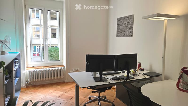 A modern, well-lit office space with a large wooden desk, dual monitors, a laptop, keyboard, mouse, and a comfortable chair. The room features large windows with a view of the street, a fan, a potted plant, and a cabinet with office supplies.