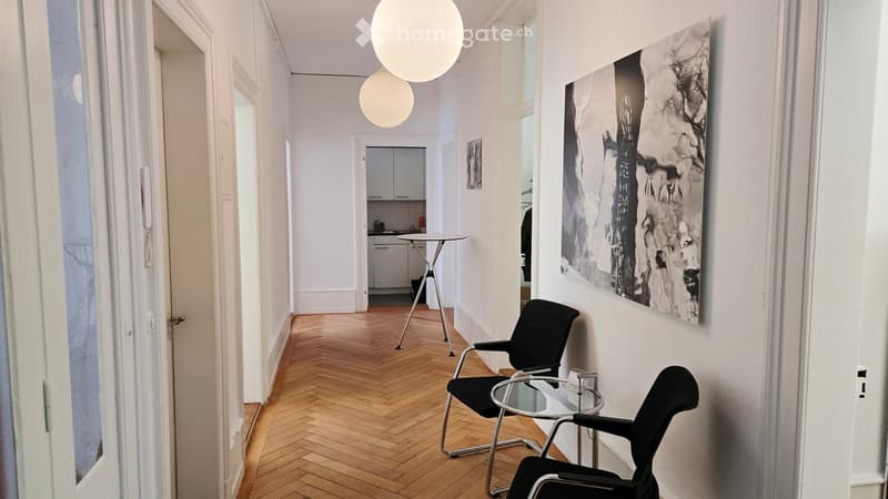 A modern, minimalist hallway with herringbone wooden flooring, white walls, and large windows. Features include a black chair, a small glass table, a stool, a cabinet, and an art piece on the wall.